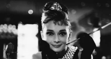 Black and white image of a woman with an updo, pearl necklace, black gloves, and holding a long cigarette holder.