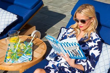 Woman in sunglasses reads a book on a blue lounge chair with lemonade on a side table.