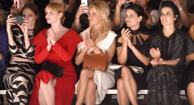 Women in stylish clothing sitting front row at a fashion show, some clapping, one taking a photo.