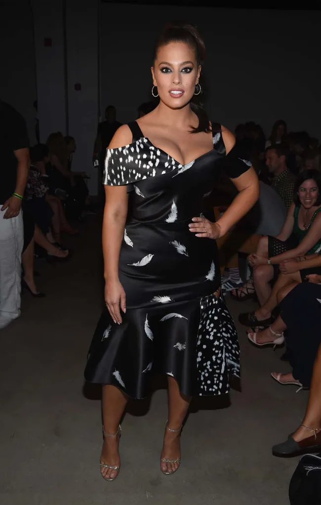 A woman in a black dress with white patterns stands confidently at a fashion event.