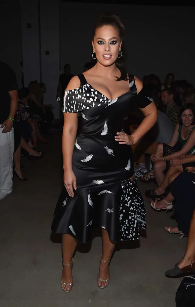 A woman in a black dress with white patterns stands confidently at a fashion event.
