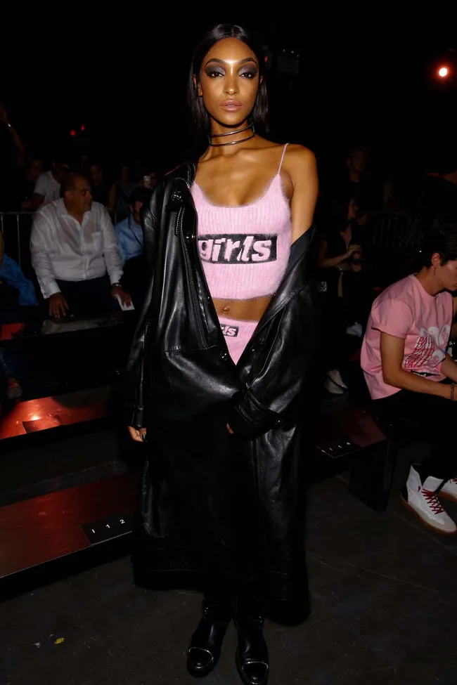 Model wears a black leather coat, pink top saying "girls," black boots, at a fashion show.
