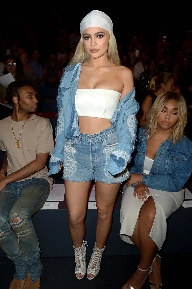 A woman wearing a white bandeau, denim jacket, and shorts stands at a fashion event, with others seated nearby.
