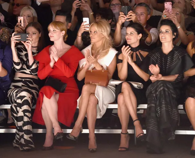 Front row of a fashion show with five women clapping and taking photos, surrounded by an audience.