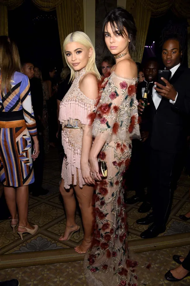 Two women in elegant dresses at a formal event, surrounded by guests.