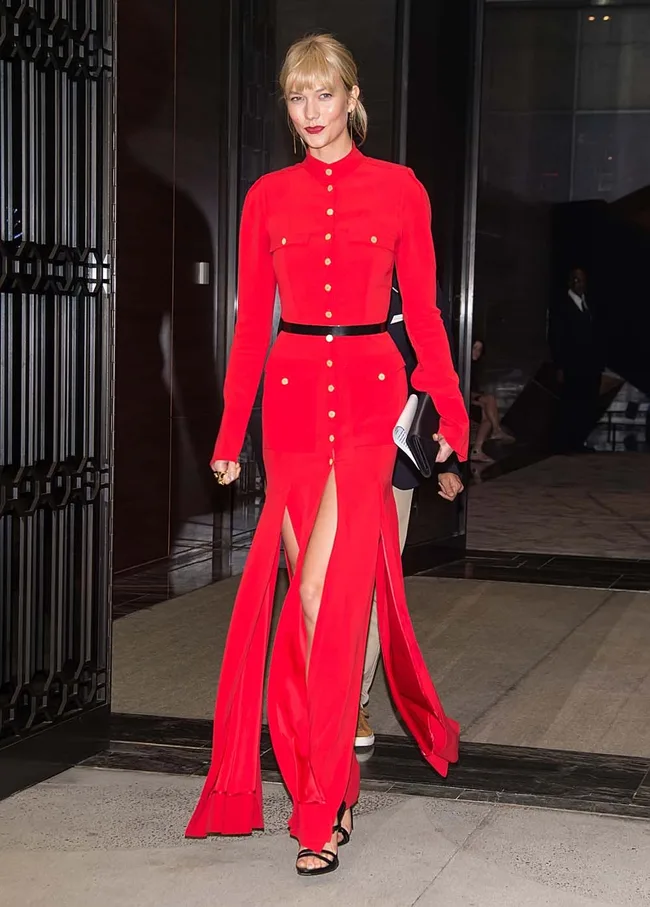 A person in a long red dress with gold buttons and high slits, walking out of a building, holding a clutch.
