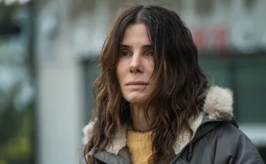 A woman with wavy brown hair in a coat with fur trim, looking solemn and reflective.