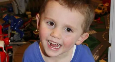 A young child smiling widely, surrounded by colorful toys in the background.