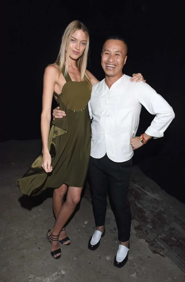 Two people pose together; one wears a green dress, and the other is in a white shirt and black pants, both smiling.
