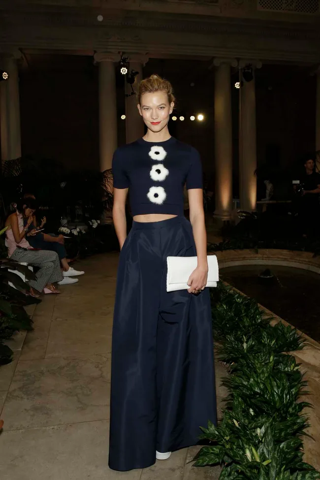 A woman in a navy outfit with white details holds a clutch at a fashionable indoor event with plants.