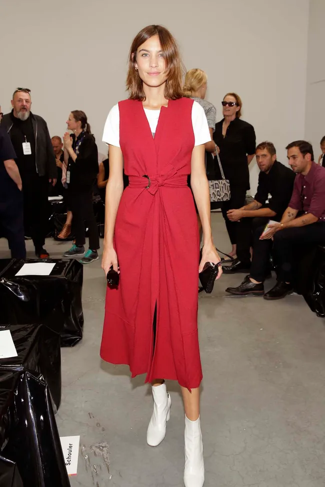 Woman in red dress with white boots at fashion event, standing and smiling, surrounded by seated attendees.