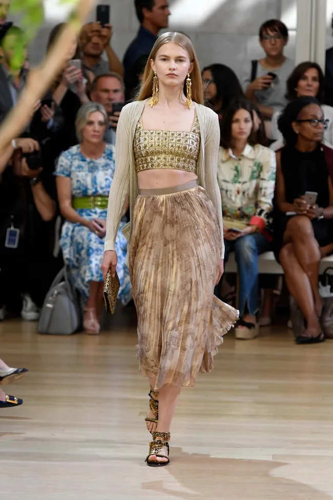 Model walks runway in patterned gold crop top, pleated skirt, cardigan, and statement earrings during a fashion show.
