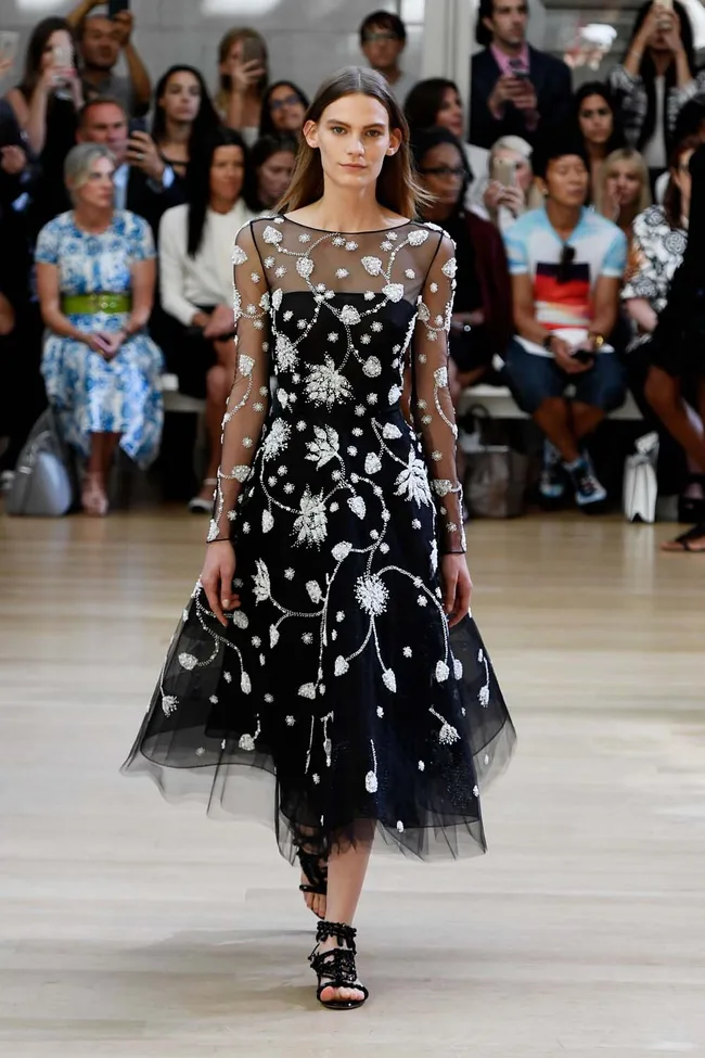 Model in a black dress with white floral patterns walks on a runway; audience seated in the background.