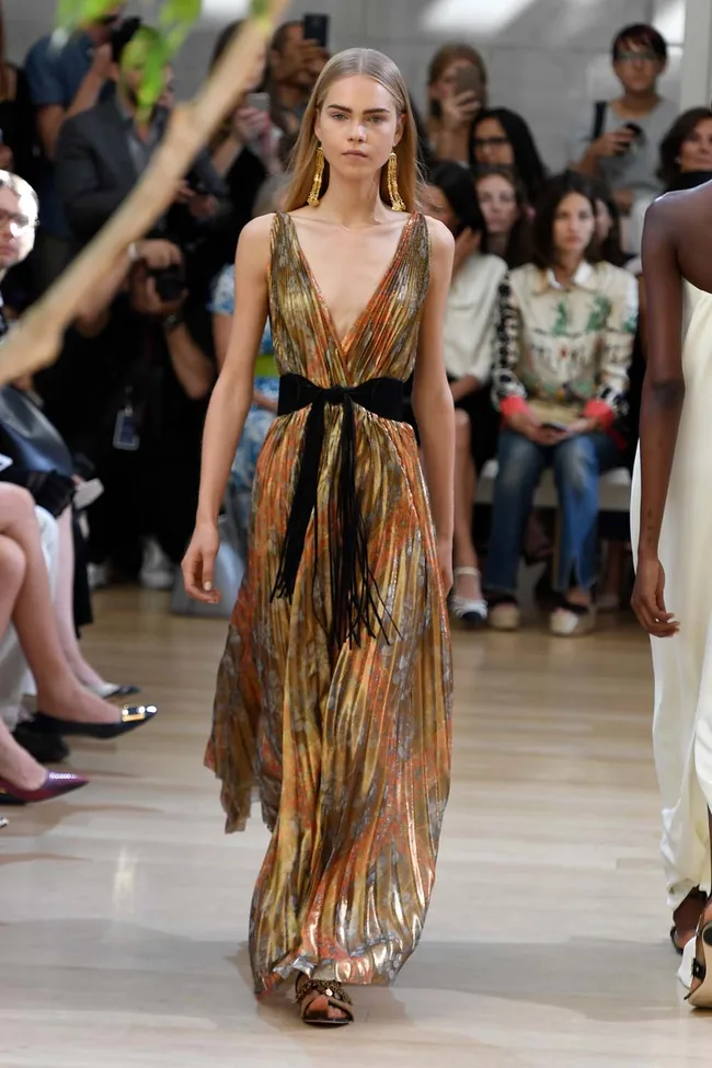 Model in a flowing, multicolored gown with black belt walking on a runway, audience in background.