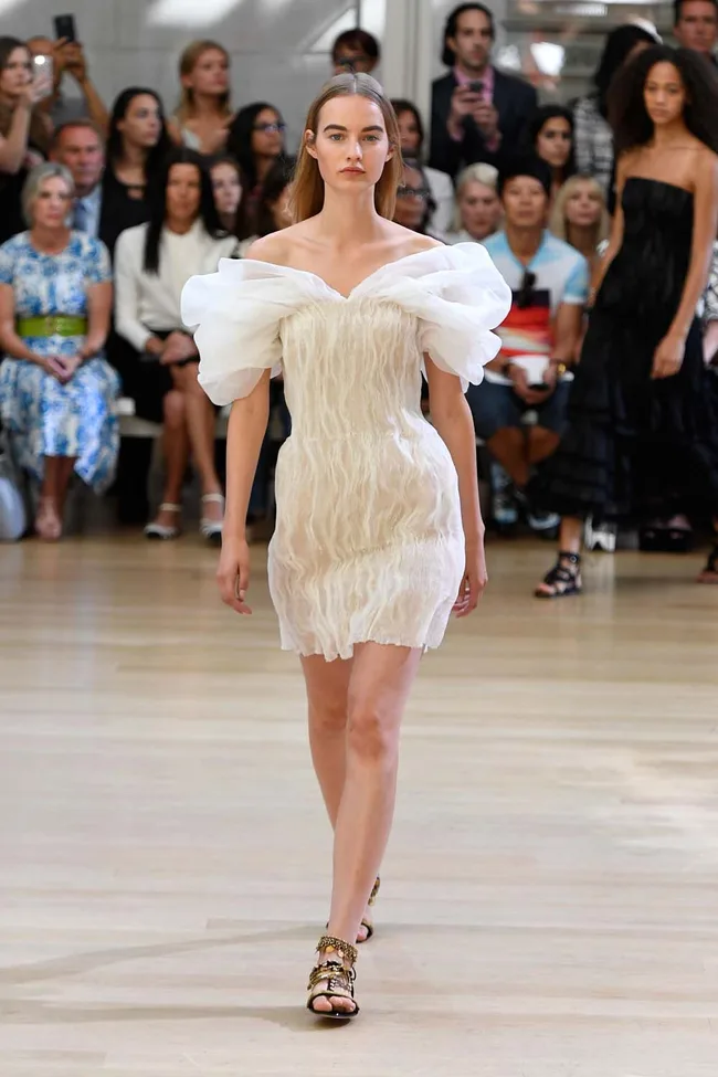 Model walking runway in a white off-shoulder dress with puff sleeves, audience seated in the background.