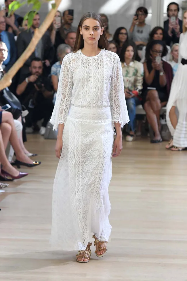 Model in a white lace dress walks down the runway, surrounded by an audience with phones and cameras.