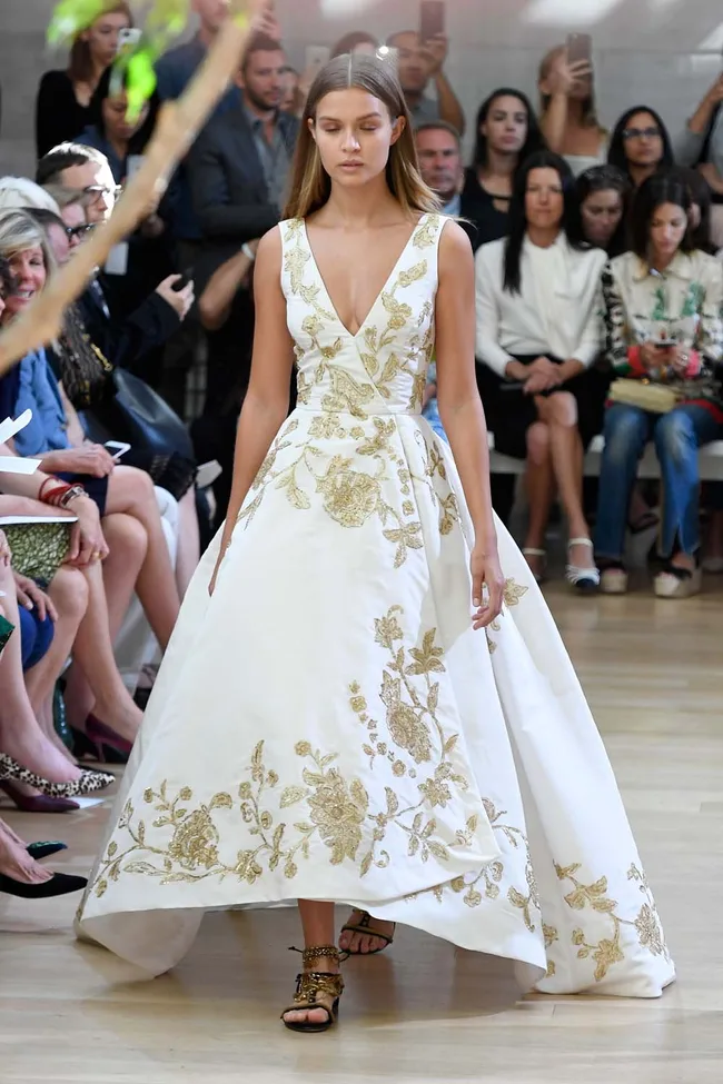 Model walking down runway wearing a white dress with gold floral embroidery, surrounded by audience.