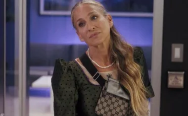 Woman with long hair, wearing a dark green dress, a pearl necklace, and a Gucci bag stands in a room with blue lighting.