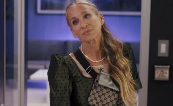 Woman with long hair, wearing a dark green dress, a pearl necklace, and a Gucci bag stands in a room with blue lighting.