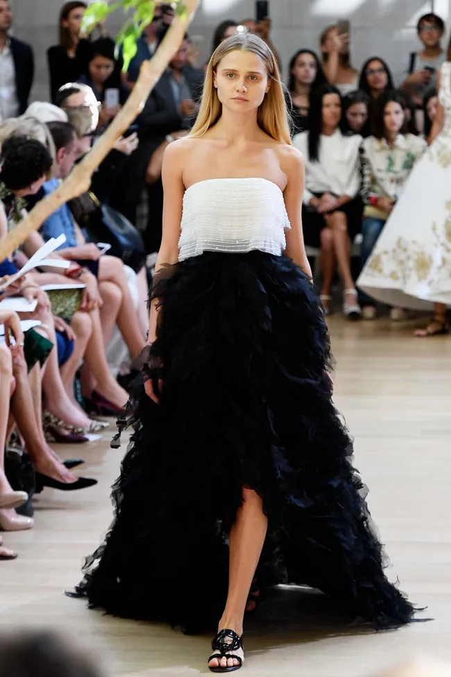 Model in strapless white and black gown walks runway at fashion show, audience in background.