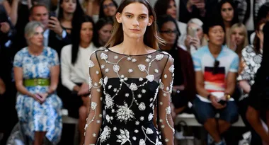 Model walks runway in a sheer black dress with white floral embellishments; audience watches in background.