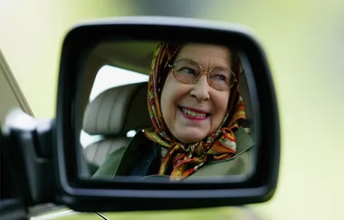 A smiling elderly woman in a headscarf is reflected in a car's side mirror.