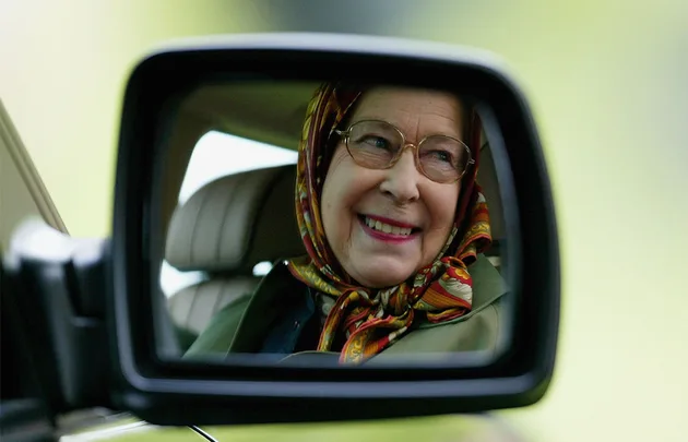 A smiling elderly woman in a headscarf is reflected in a car's side mirror.