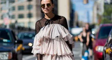 Woman in polka dot ruffled dress and sunglasses walking on a city street with cars and buildings in the background.