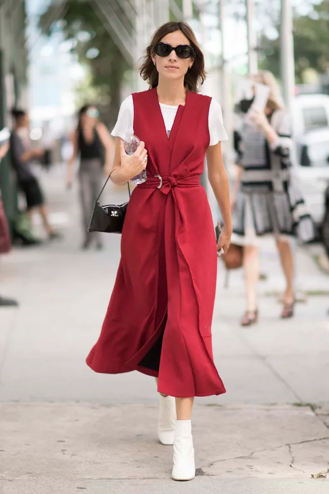Woman in a red dress and white boots walking on a street, holding a handbag and water bottle, wearing sunglasses.