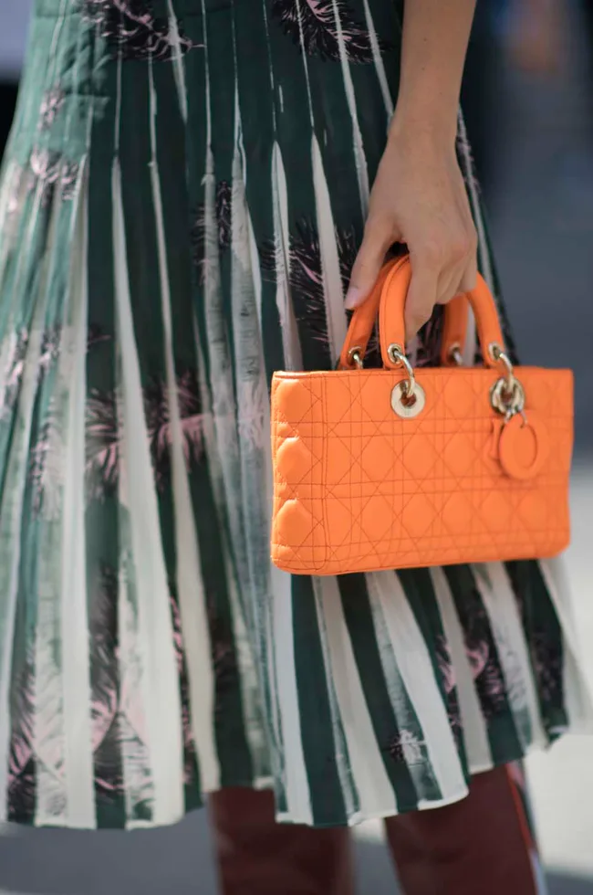 Woman holding an orange quilted handbag, wearing a green and white pleated skirt with abstract patterns.