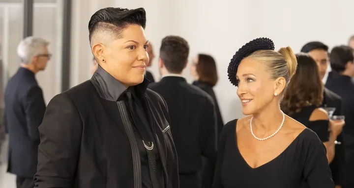 Two well-dressed individuals conversing at a formal event with other attendees in the background.