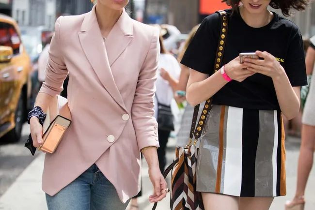 Two people walking, one in a pink blazer holding a phone, the other in a striped skirt holding a smartphone, city street scene.