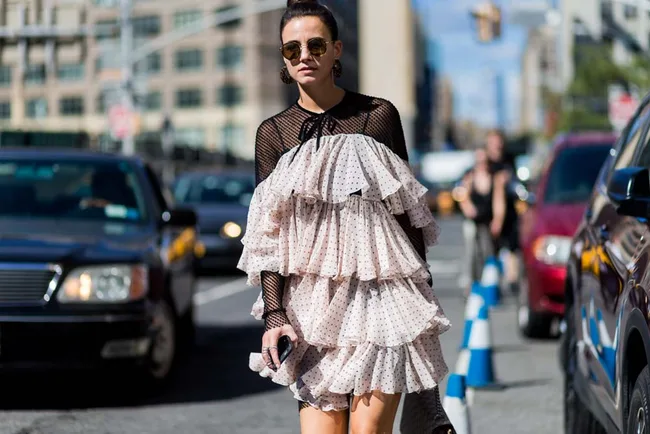 Woman in polka-dot ruffled dress and sunglasses walking on a city street with cars in the background.