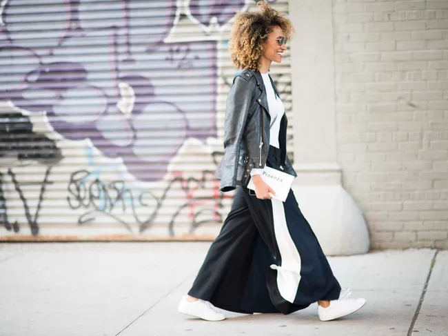 Woman in a leather jacket and long skirt walking past graffiti-covered wall, holding a "Proenza" book.