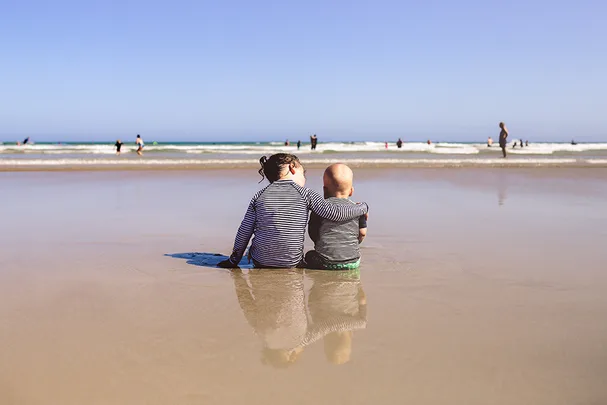 Children in striped shirts sit embracing on a beach, looking at the ocean with people playing in the water.