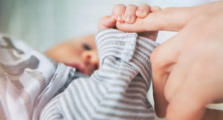 Baby in a striped outfit gripping an adult's finger gently.