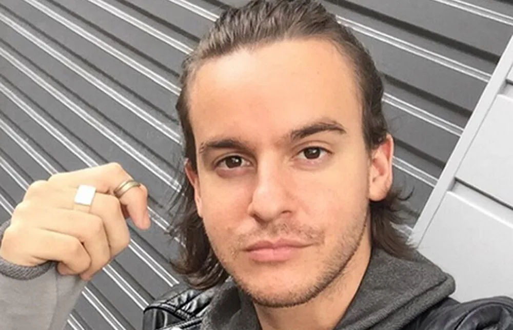 Young man with long hair tied back, wearing a leather jacket and rings, standing in front of a metal shutter.