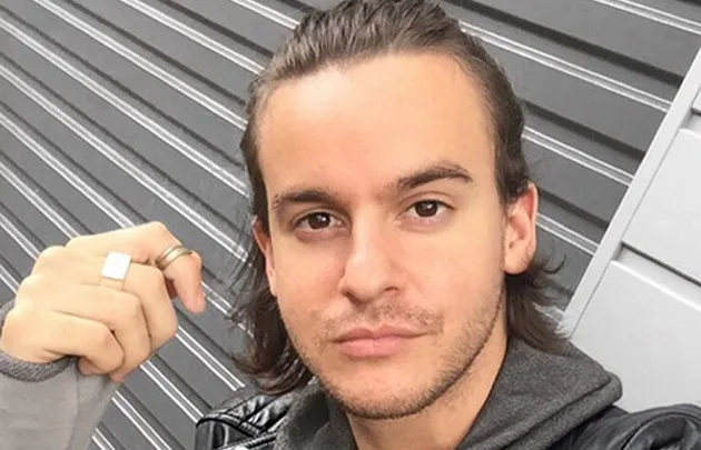 Young man with long hair tied back, wearing a leather jacket and rings, standing in front of a metal shutter.