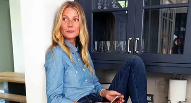 Woman with long blonde hair in denim shirt and jeans sits on kitchen counter holding a phone.