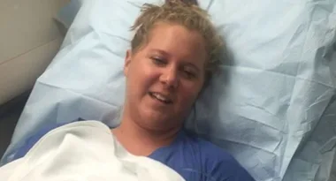 Person smiling while lying in a hospital bed, covered with a light blue sheet.