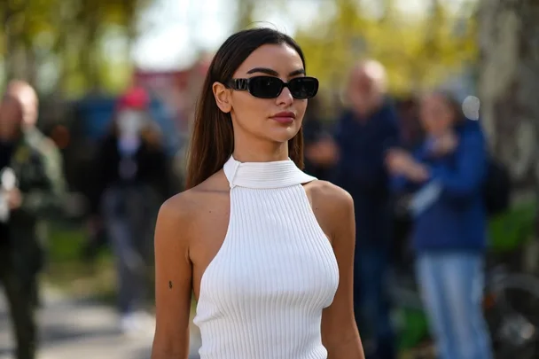 Woman in a white sleeveless ribbed top, wearing black sunglasses, with blurred crowd and trees in the background.