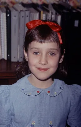Young girl with short brown hair and a red bow, wearing a blue dress with embroidered collar, smiling in front of bookshelves.