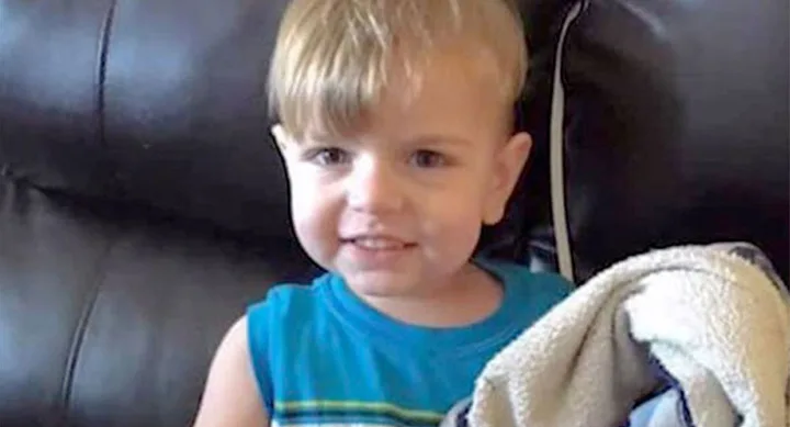 A young child with blond hair wearing a blue shirt, sitting on a couch and holding a blanket, smiling at the camera.