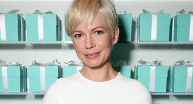 A person with short blonde hair wearing a white top stands in front of a shelf with blue gift boxes.