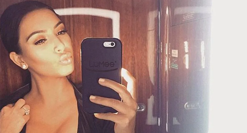 Woman taking a selfie in a mirror with a smartphone, making a pouty face, in a dimly lit room.