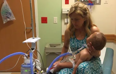 Woman holding a child connected to medical equipment in a hospital room.