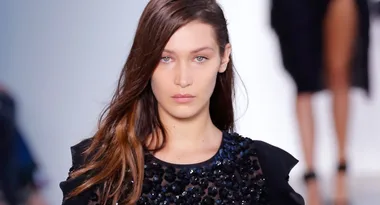 Young woman with long hair walks runway in a black embellished dress.