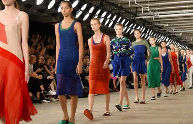 Models walk down a runway wearing colorful dresses at a fashion show, with an audience watching.