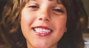 A child with light brown hair smiles at the camera, showing their teeth.