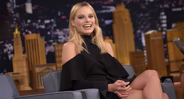 A woman with blonde hair in a black dress smiles while sitting on a talk show set with a cityscape backdrop.